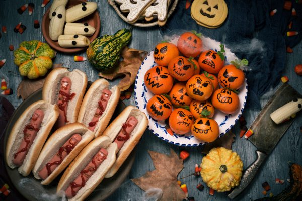 high-angle shot of some funny halloween food, such as tangerines as carved pumpkins with funny faces, bananas with eyes as ghosts or hotdogs in the shape of bloody fingers, on a rustic table