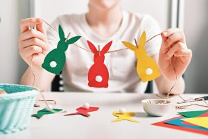 DIY Easter home decor. Happy boy make garland of colored paper, twine and pom-poms. Selective focus.