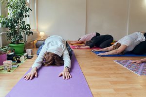 A group of senior women engage in various yoga exercises, including neck, back, and leg stretches, under the guidance of a trainer in a sunlit space, promoting well-being and harmony.